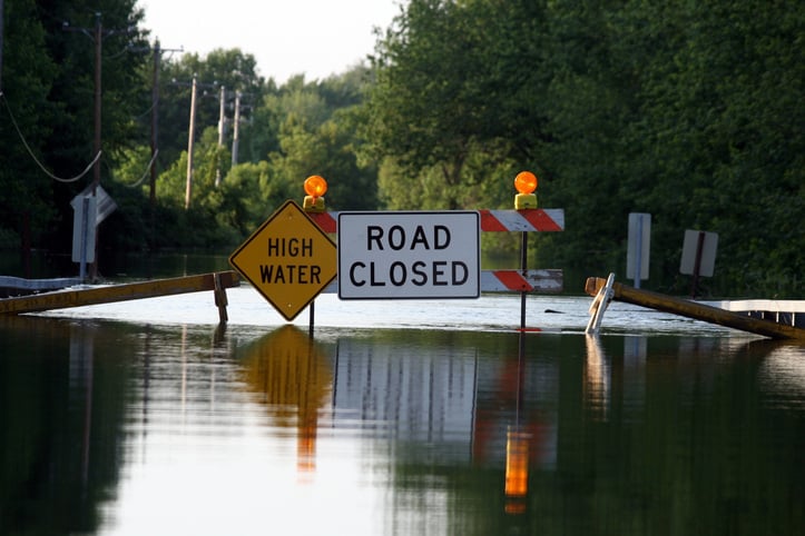 Flooded road due to a natural disaster with ‘Road Closed’ and ‘High Water’ signs.